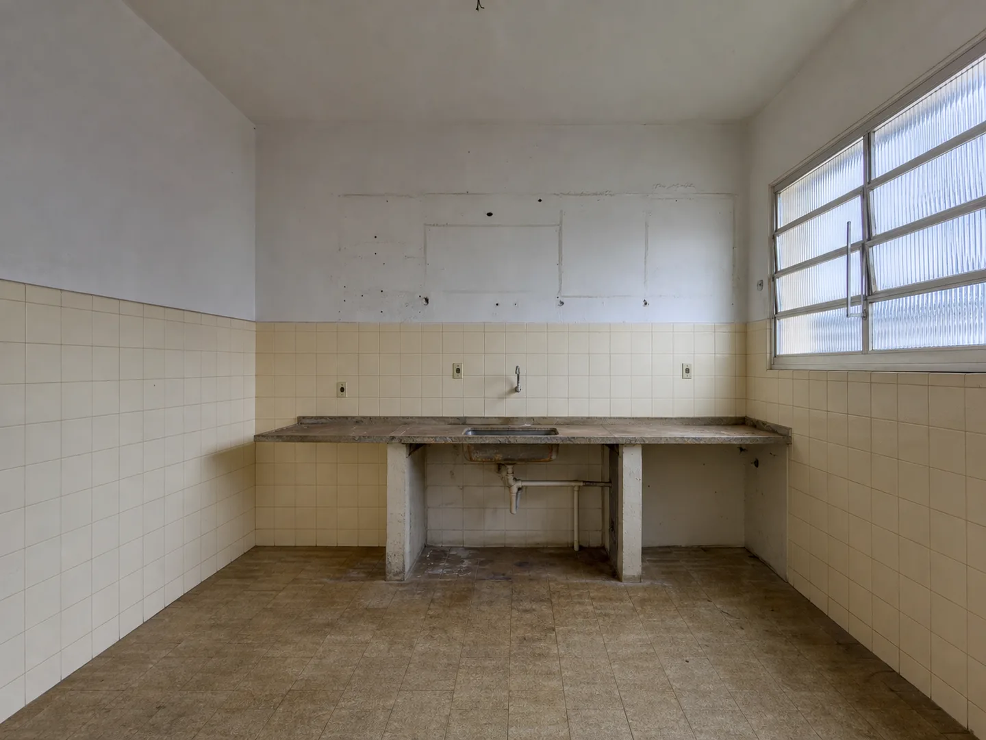Cozinha vazia antes da reforma, com azulejo antigo até meia altura, bancada desgastada e hidráulica aparente.